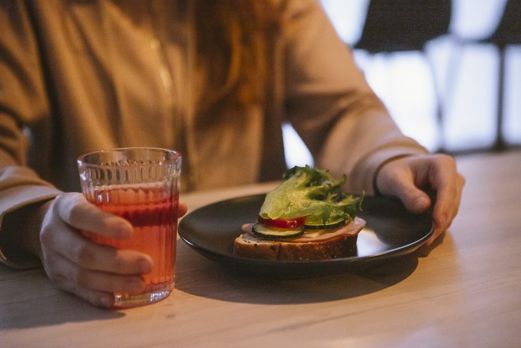 Henkilö istuu pöydän ääressä voileipä ja juomalasi edessään.